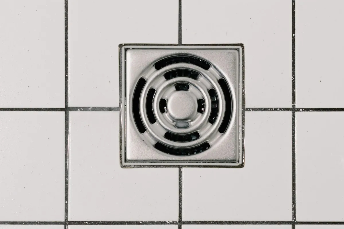Siphon de sol carré en inox encastré dans un carrelage blanc de salle de bain.